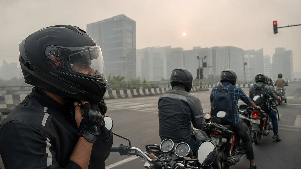 A queue of two-wheeler riders waiting at a signal on a Noida flyover, glass office towers softened by afternoon smog haze behind them.