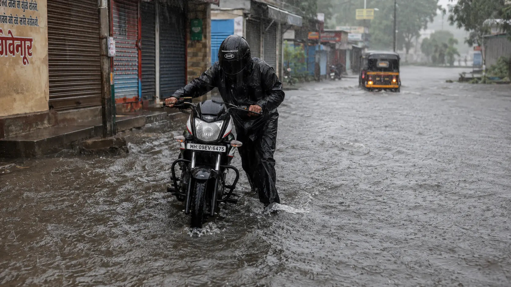 A rider in rain gear pushing a motorcycle through ankle-deep water on a flooded Kolhapur street under heavy grey monsoon skies.