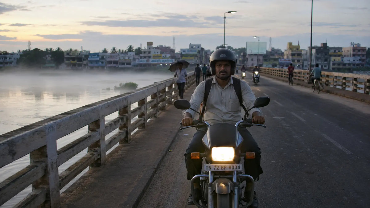 A rider crossing the river bridge between Tirunelveli and Palayamkottai at sunrise, mist rising off the Thamirabarani, helmet strap loose under his chin.