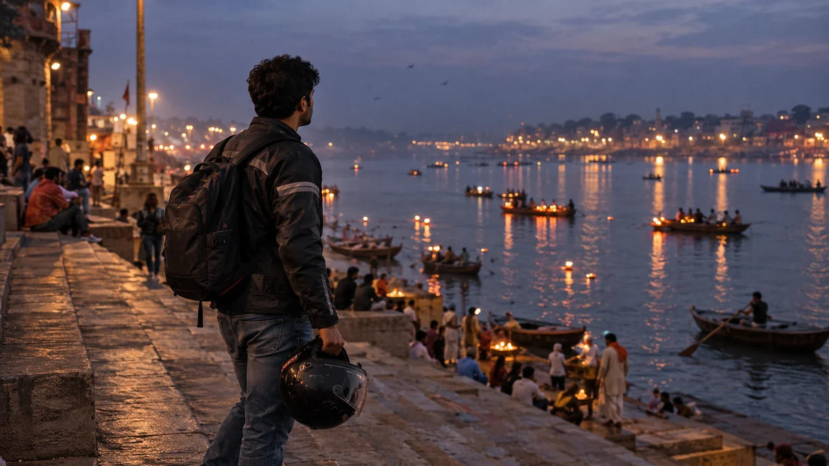 A rider pauses on a wide stone ghat at dusk, helmet held at his side, distant diya flames flickering along the Ganga behind him.