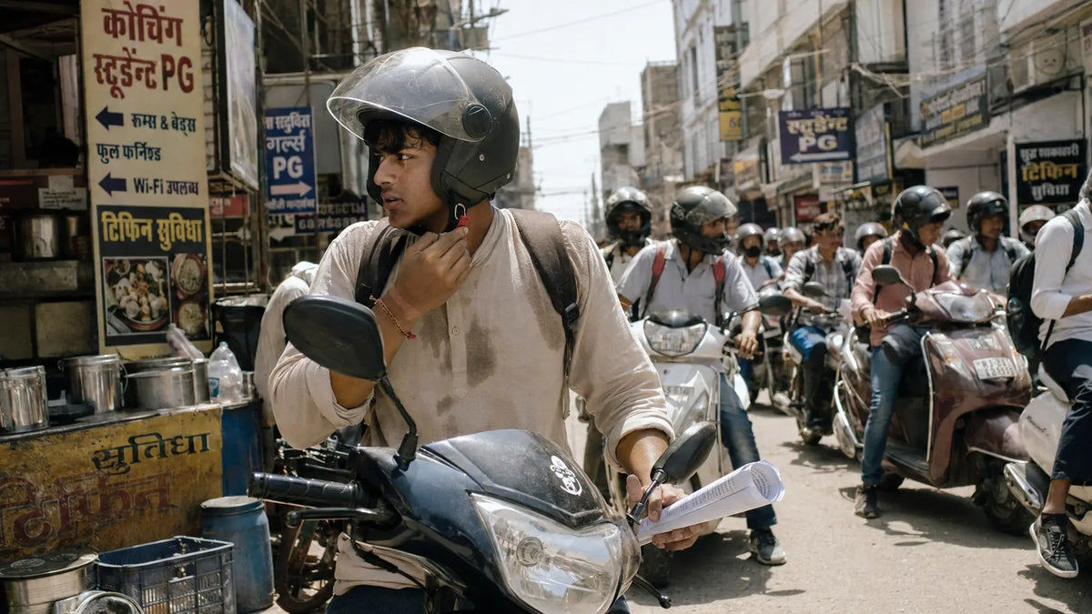 A student rider fastens his helmet strap outside a Kota coaching center gate at midday, scooters parked bumper to bumper along the lane in harsh, glaring sunlight.