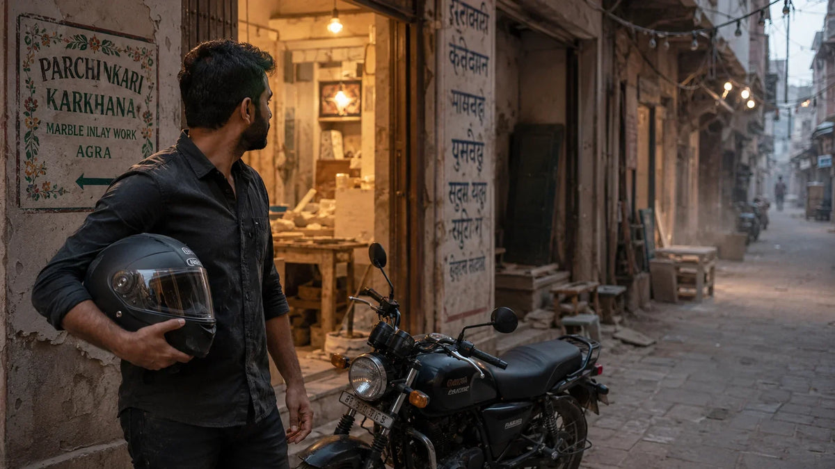 A rider pauses his motorcycle in a narrow Agra marble-inlay artisan lane at dusk, helmet resting under one arm, dust catching the fading light.
