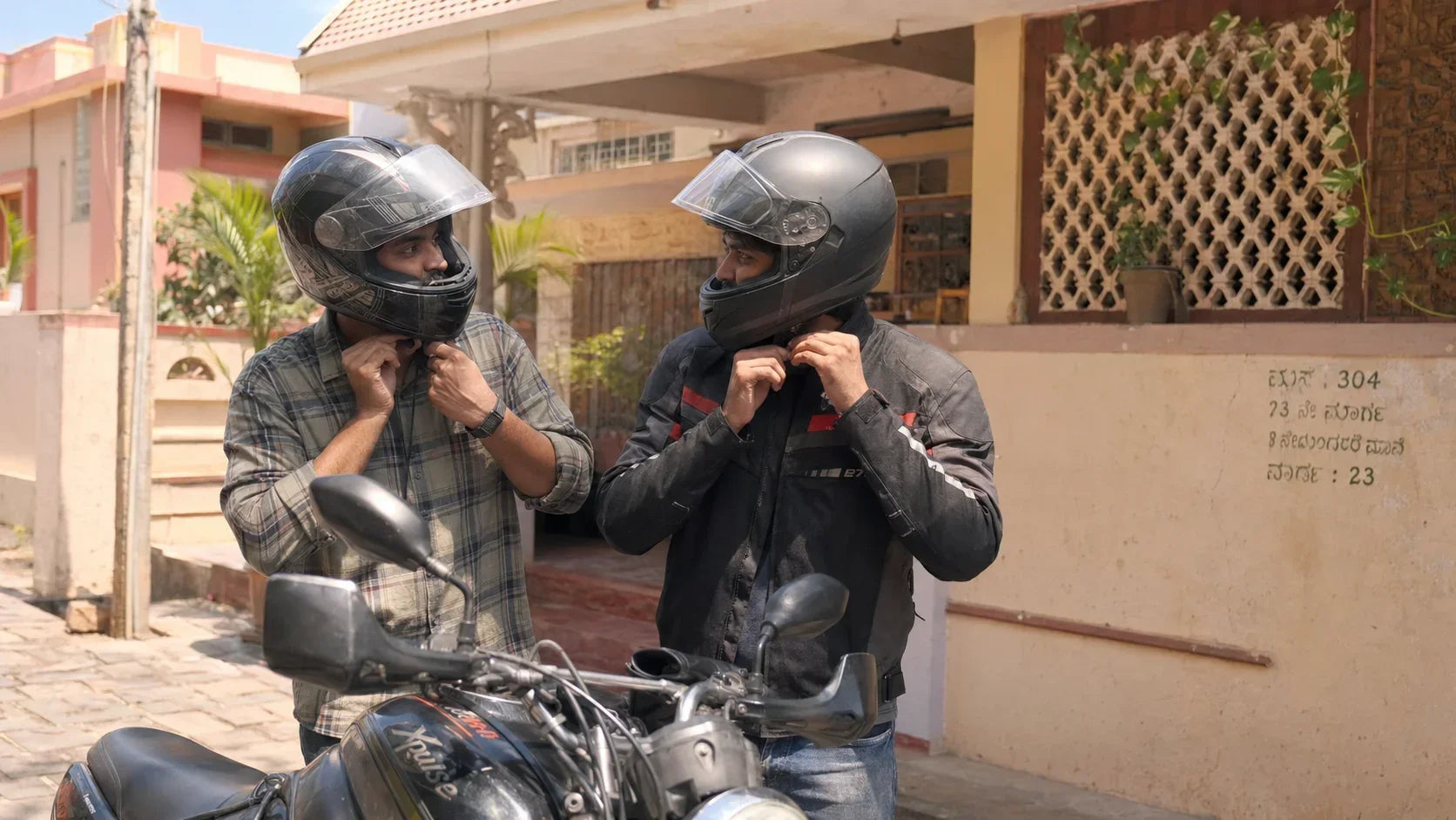 Two riders adjusting helmet straps together in a sunlit Mysuru courtyard lane at midday.
