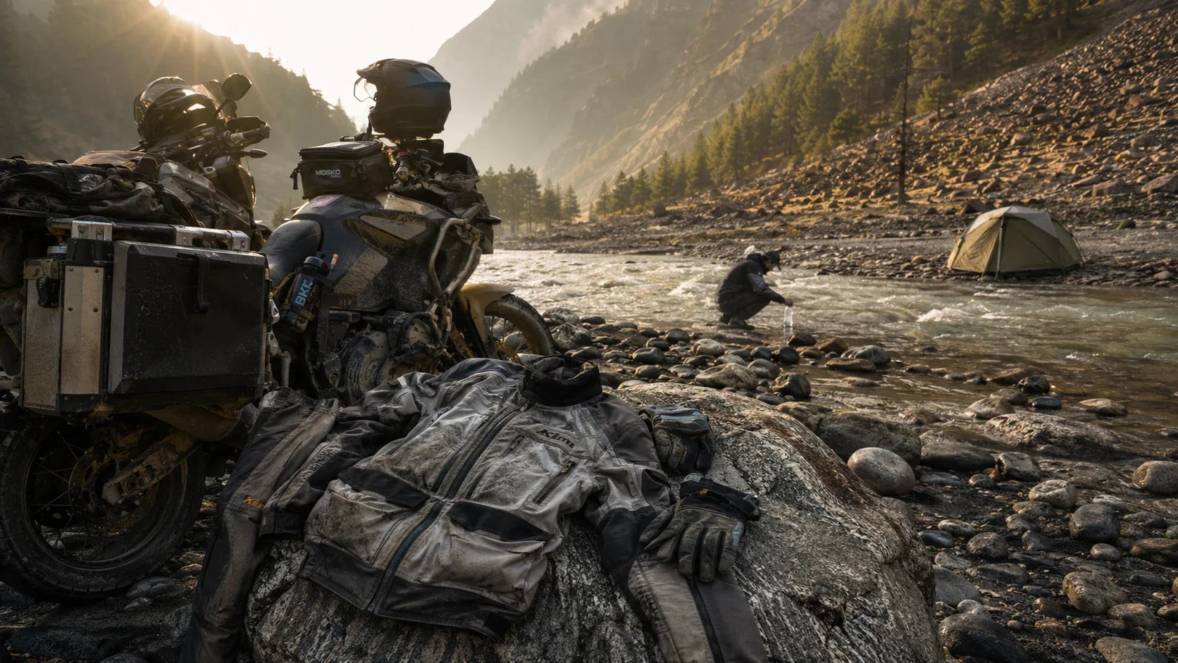 A motorcycle parked beside a Himalayan river at dawn, panniers open and riding gear airing on a rock near a small tent.