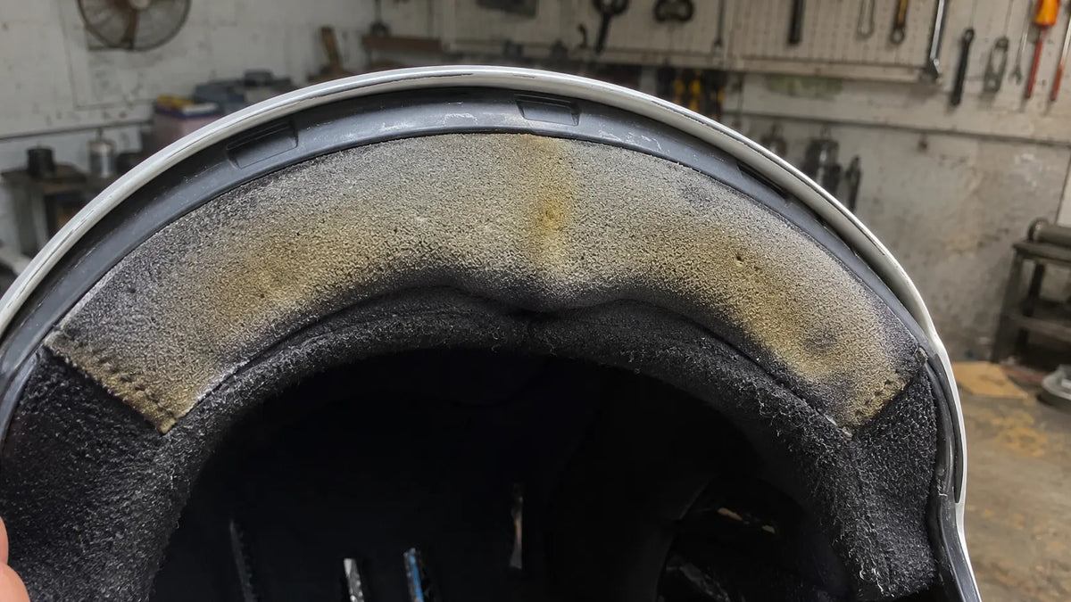 Extreme close-up of a motorcycle helmet's foam liner in a home garage under harsh midday light, showing discoloration at the brow pad.
