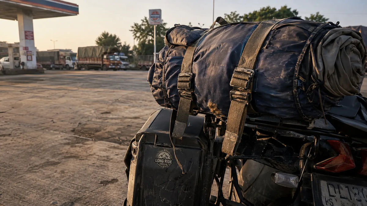 A packed motorcycle pannier and rolled sleeping bag strapped to a bike at a roadside stop during golden hour, dust visible on the gear.