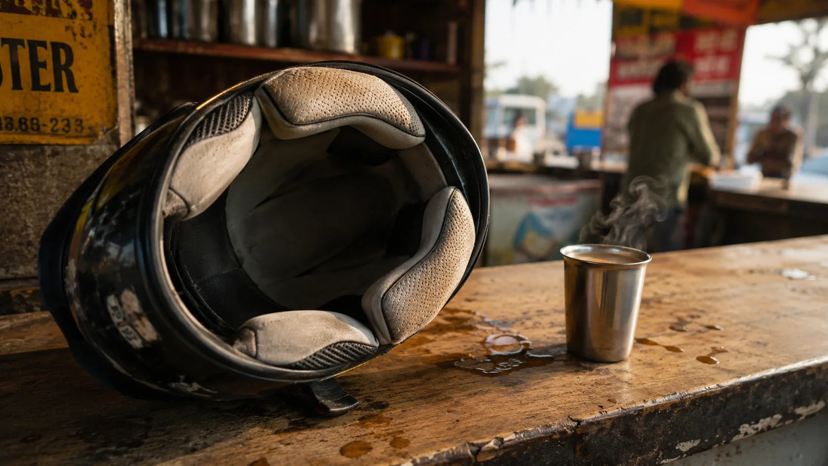 Close-up of a well-used motorcycle helmet resting on a roadside dhaba counter, foam liner showing faint discoloration at the brow pad.