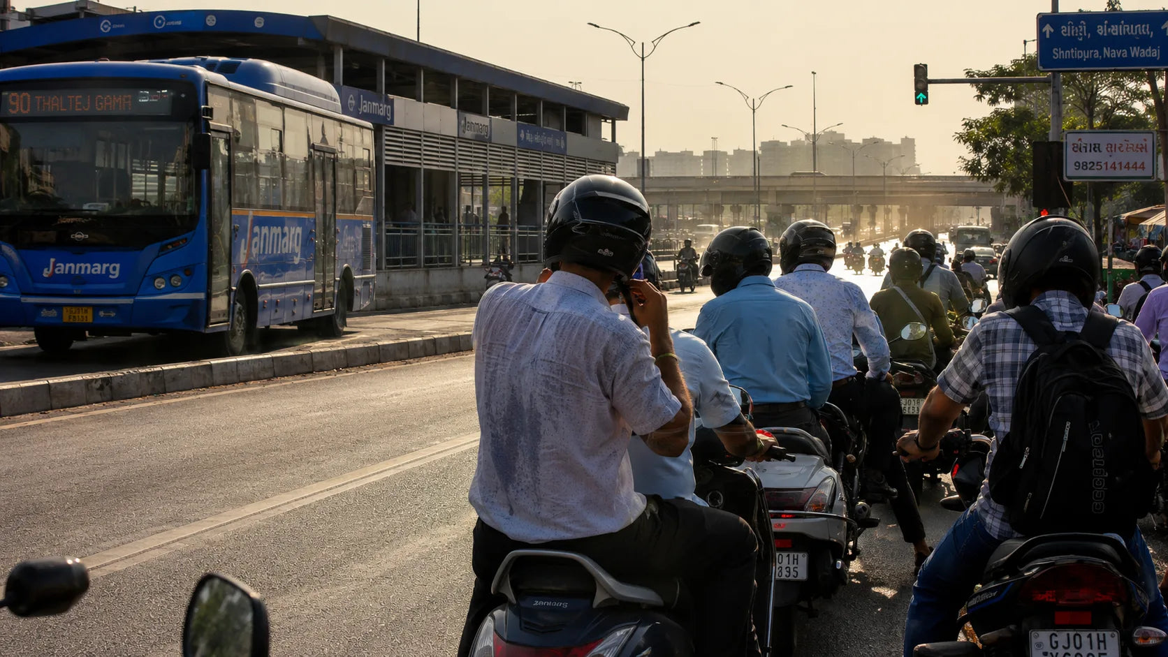 A cluster of two-wheeler riders wait beside a blue Janmarg BRTS bus at a signal on an Ahmedabad flyover under harsh afternoon sun, heat haze rising off the road.