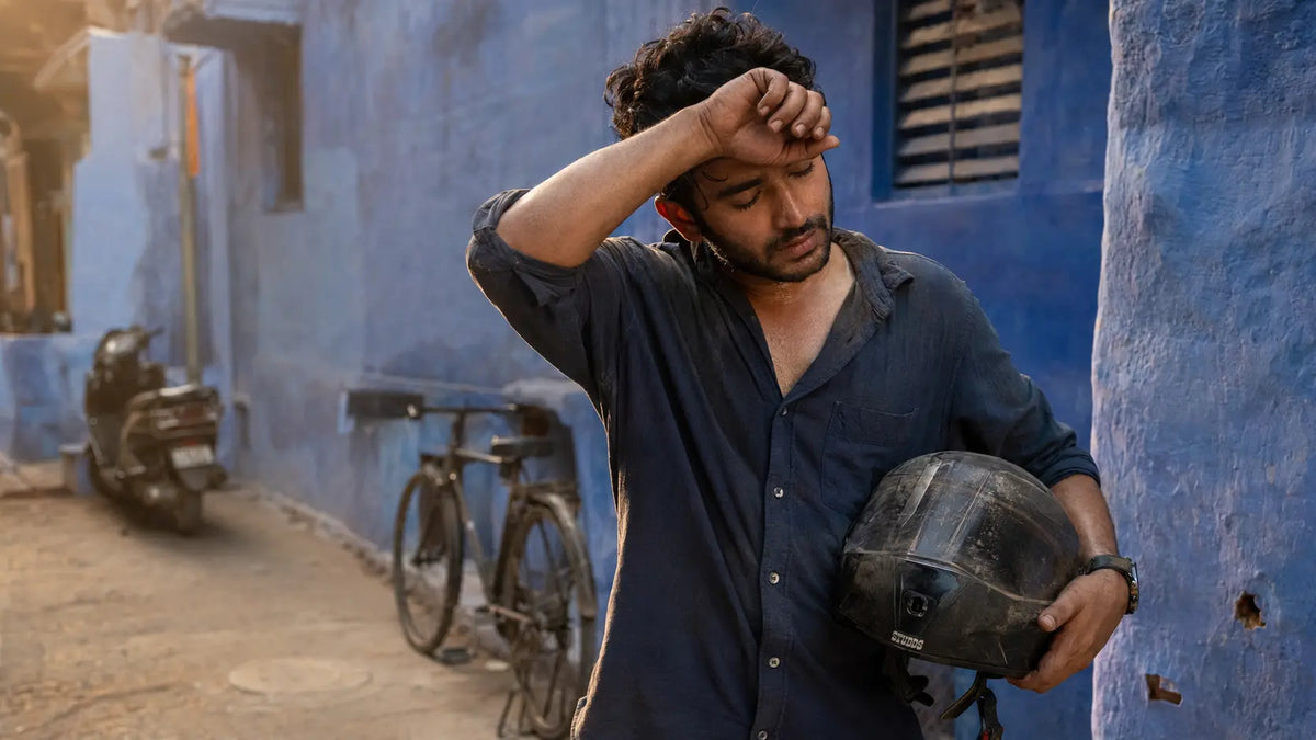 A rider stands in a narrow blue-painted lane in Jodhpur's old city in warm late-afternoon light, helmet under one arm, wiping sweat and dust from his brow.