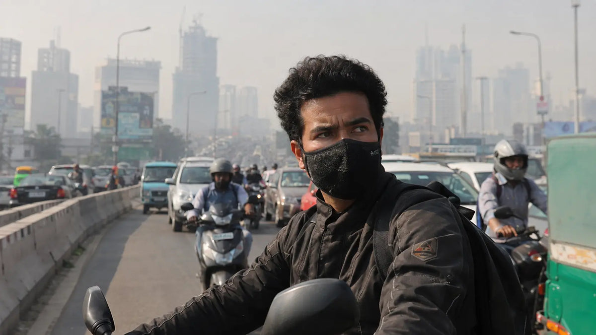 A rider wearing a black anti-pollution mask rides through hazy midday city traffic, smog visible behind the skyline.