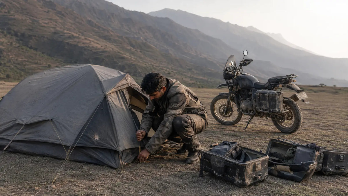 A rider crouches beside a partially pitched tent next to a parked motorcycle in late-afternoon light in a highland valley, panniers open on the ground.