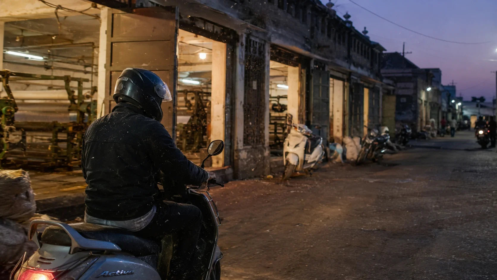 A rider in a full-face helmet on a scooter passing rows of powerloom sheds in Solapur at dusk, cotton lint drifting through the shed light.