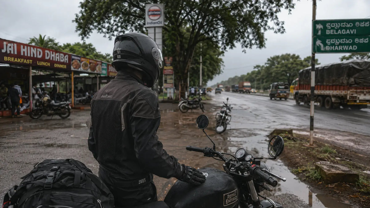 A rider pausing at a fuel stop on the NH48 highway outside Belgaum at midday, cantonment-era trees lining the road behind the parked motorcycle.