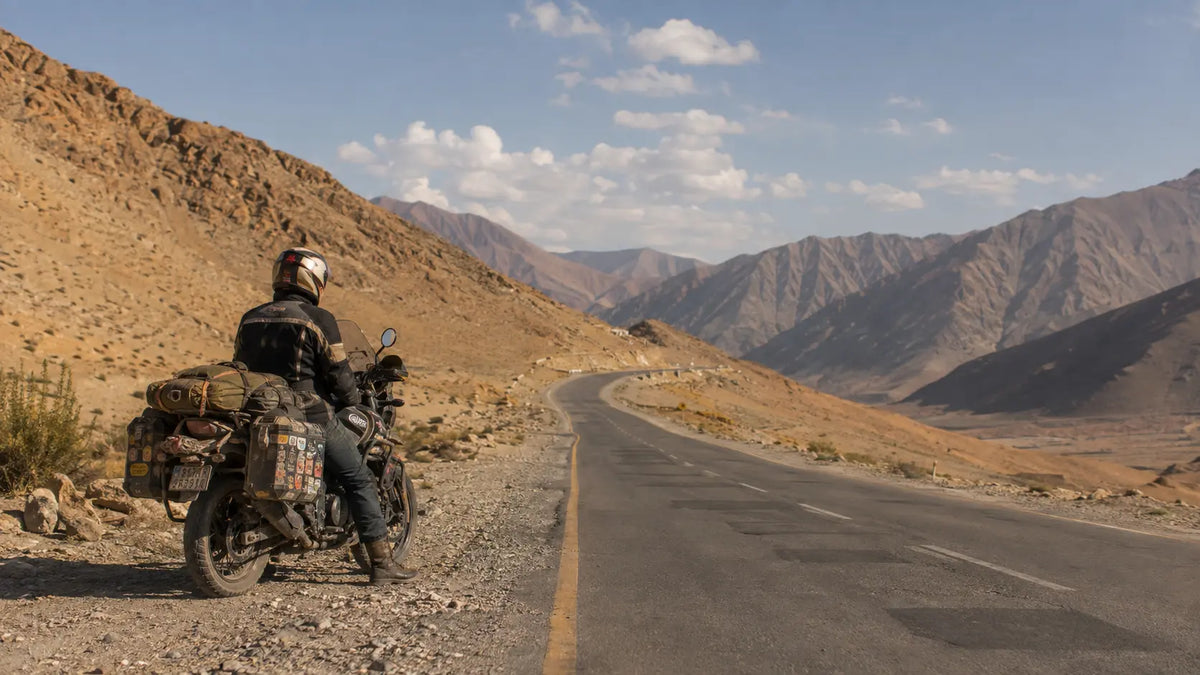 A lone rider stopped on an empty highway shoulder in late-afternoon light, looking out toward hills with the bike parked beside them and no other traffic in sight.