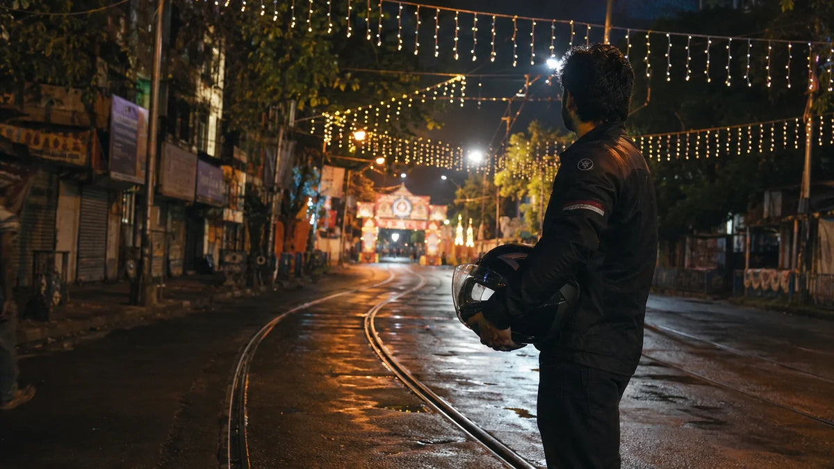 A solo rider pauses beside old tram tracks on a Kolkata street lit by strings of Durga Puja pandal lights at night, helmet held under one arm.