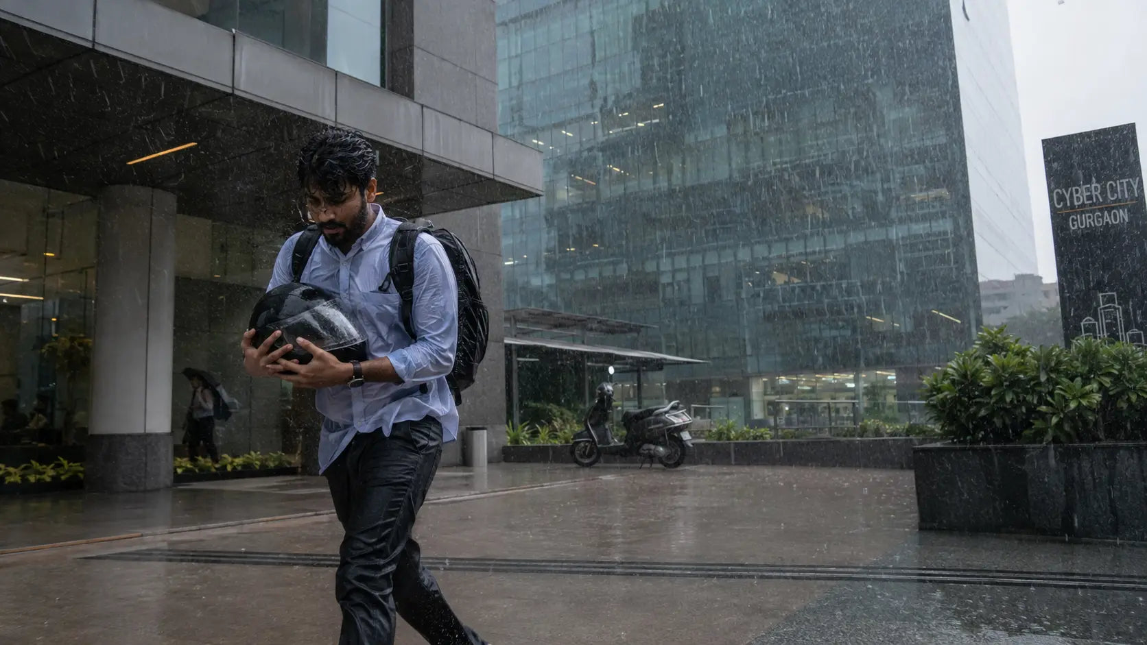 Rider in office wear hurrying toward a Cyber City tower portico with a helmet clutched to their chest as a sudden monsoon downpour streaks past the glass facade behind them.