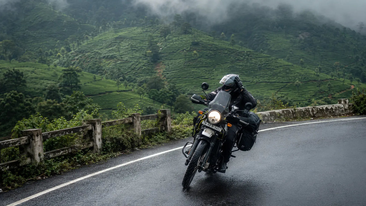A rider climbing a hairpin ghat road toward Valparai steadies his helmet against a gust of wind, tea estates and fast-moving monsoon clouds in the background.