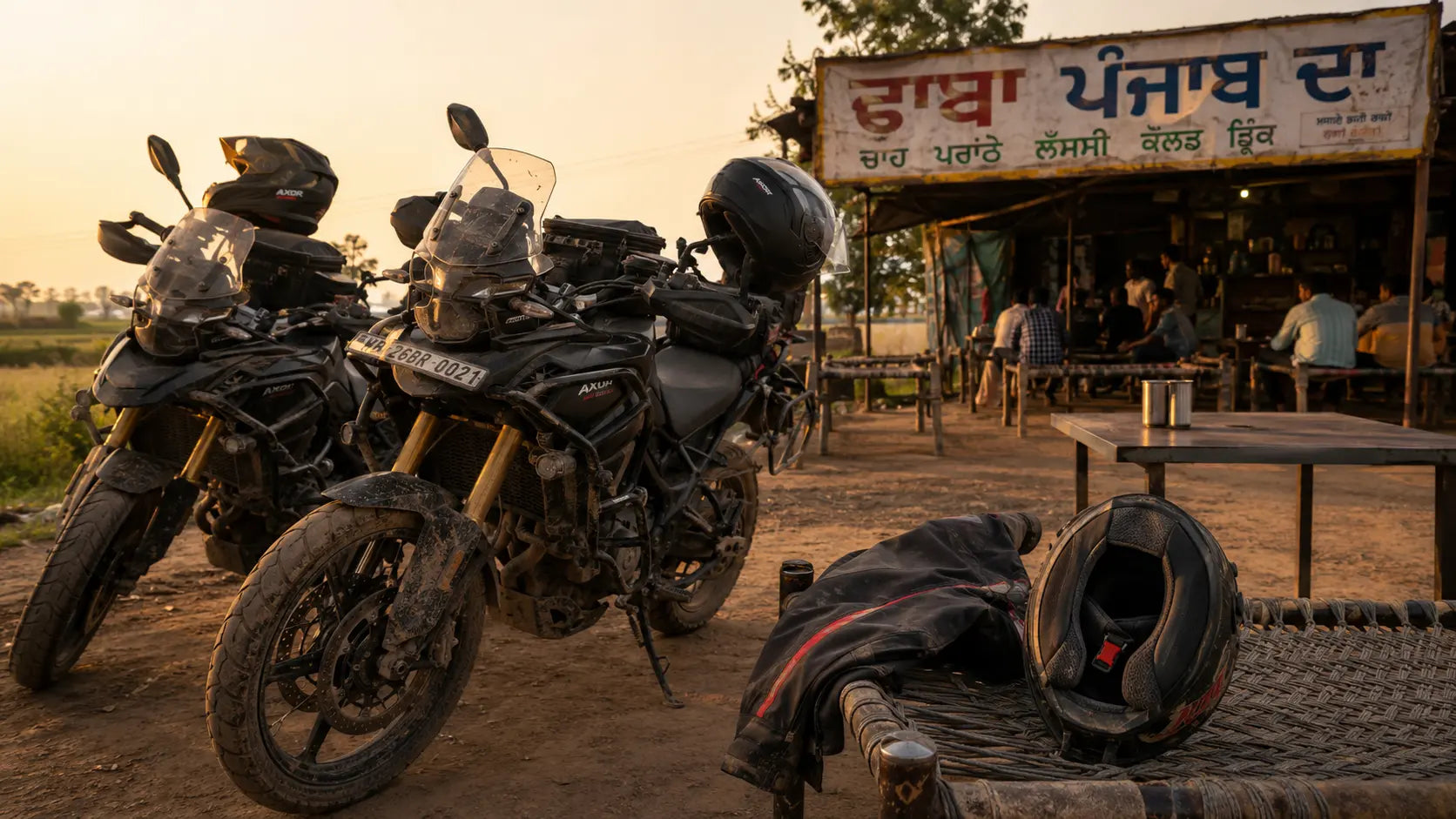 Two touring motorcycles parked outside a roadside Punjab dhaba at golden hour, charpais and steel plates on the table, dust hanging in the warm light.