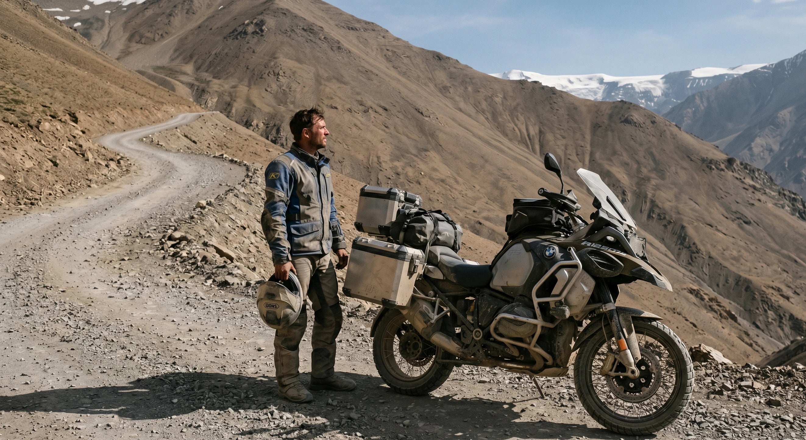 A solo rider on a loaded motorcycle crossing a dusty high-altitude pass in Spiti Valley, snow peaks in the background