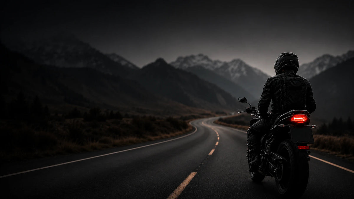 A person riding a black motorcycle on a winding road with dark mountains in the background under a gloomy sky.